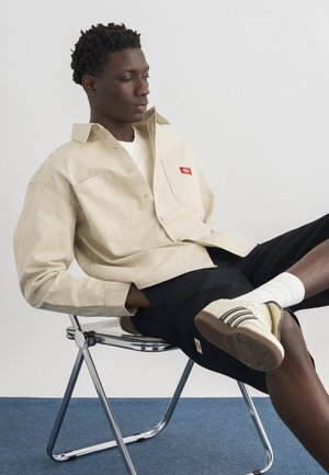 Homme en chemise rayée beige et short noir assis sur une chaise en métal, une jambe croisée, portant des chaussettes blanches et des baskets beiges à rayures noires.
