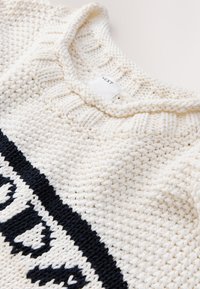 Suéter de punto color crema con cuello redondo acanalado. Presenta un patrón gráfico azul marino en la parte inferior y detalles texturizados de trenzado.