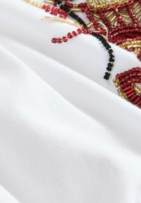 Țesătură albă cu textură, decorată cu mărgele roșii, negre și aurii într-un model floral, evidențiind detalii intricate de broderie.