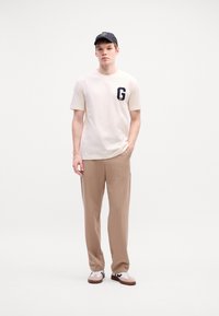 Jeune homme portant un t-shirt blanc avec un logo "G" noir, un pantalon beige, une casquette noire et des baskets blanches, se tenant devant un fond blanc uni.