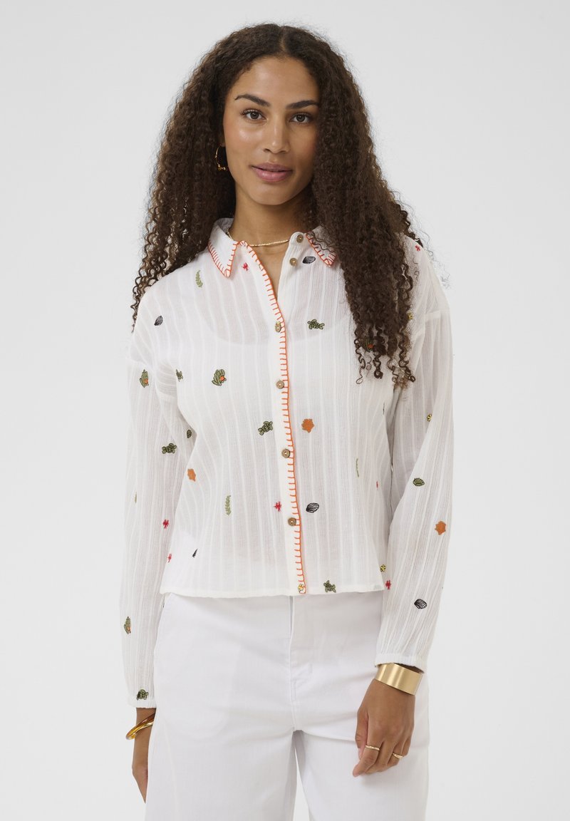 Femme aux longs cheveux bouclés, portant une chemise blanche à boutons avec de petites broderies colorées et un pantalon blanc, posant devant un fond uni.