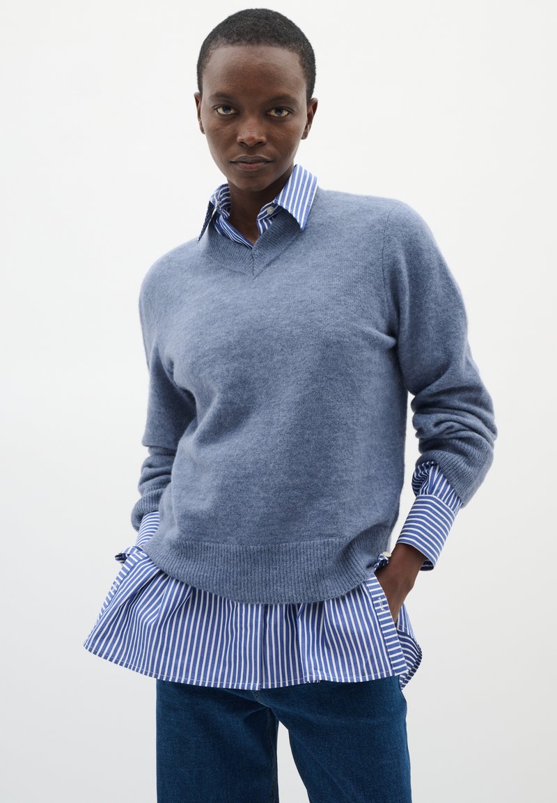 Svetlo modra volnena pulover s V-izrezom, nadeljen na modro-belo prugasto srajco s nabranim robom, v kombinaciji z jeans hlačami.