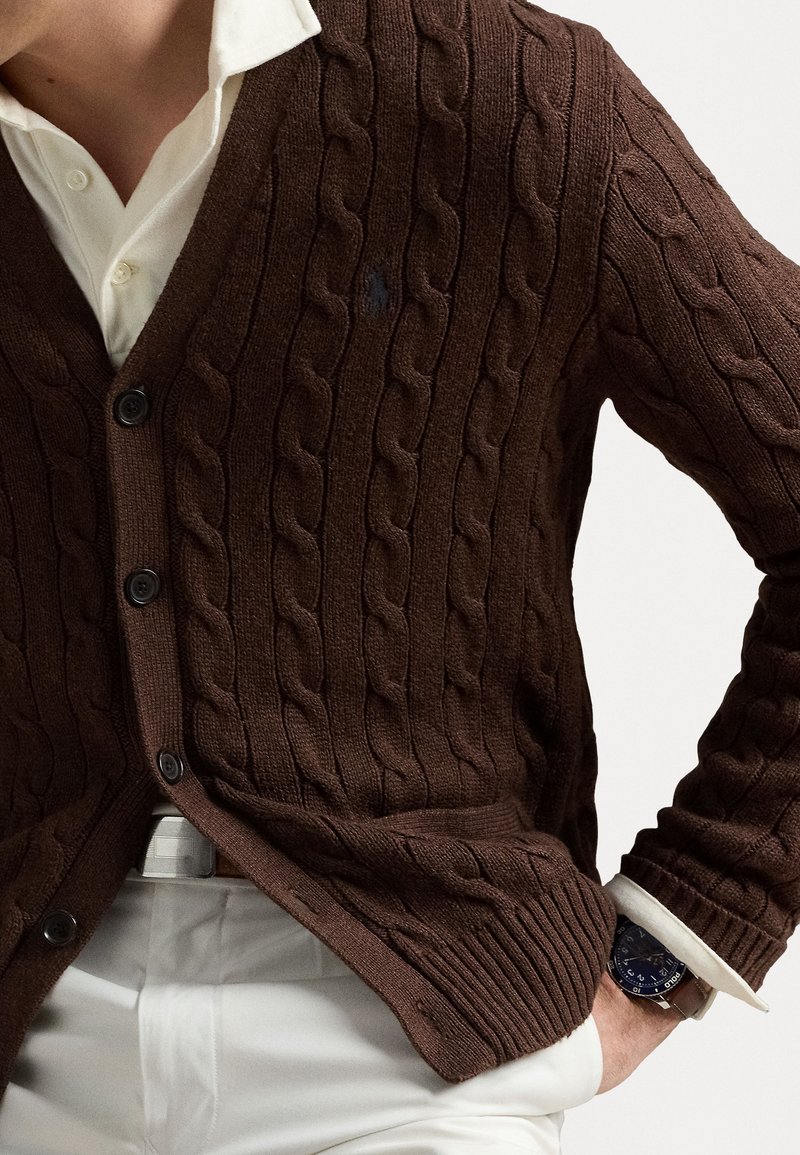 Cardigan marrone a maglia con motivi a treccia, chiusura a bottoni e polsini a costine. Abbinato a una camicia bianca e pantaloni chiari.