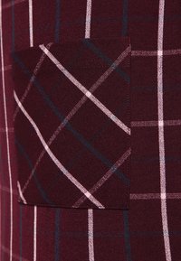 Tela a cuadros burdeos con un bolsillo en el pecho, con líneas blancas y azul marino formando un patrón de cuadrícula. Textura suave y diseño estructurado.