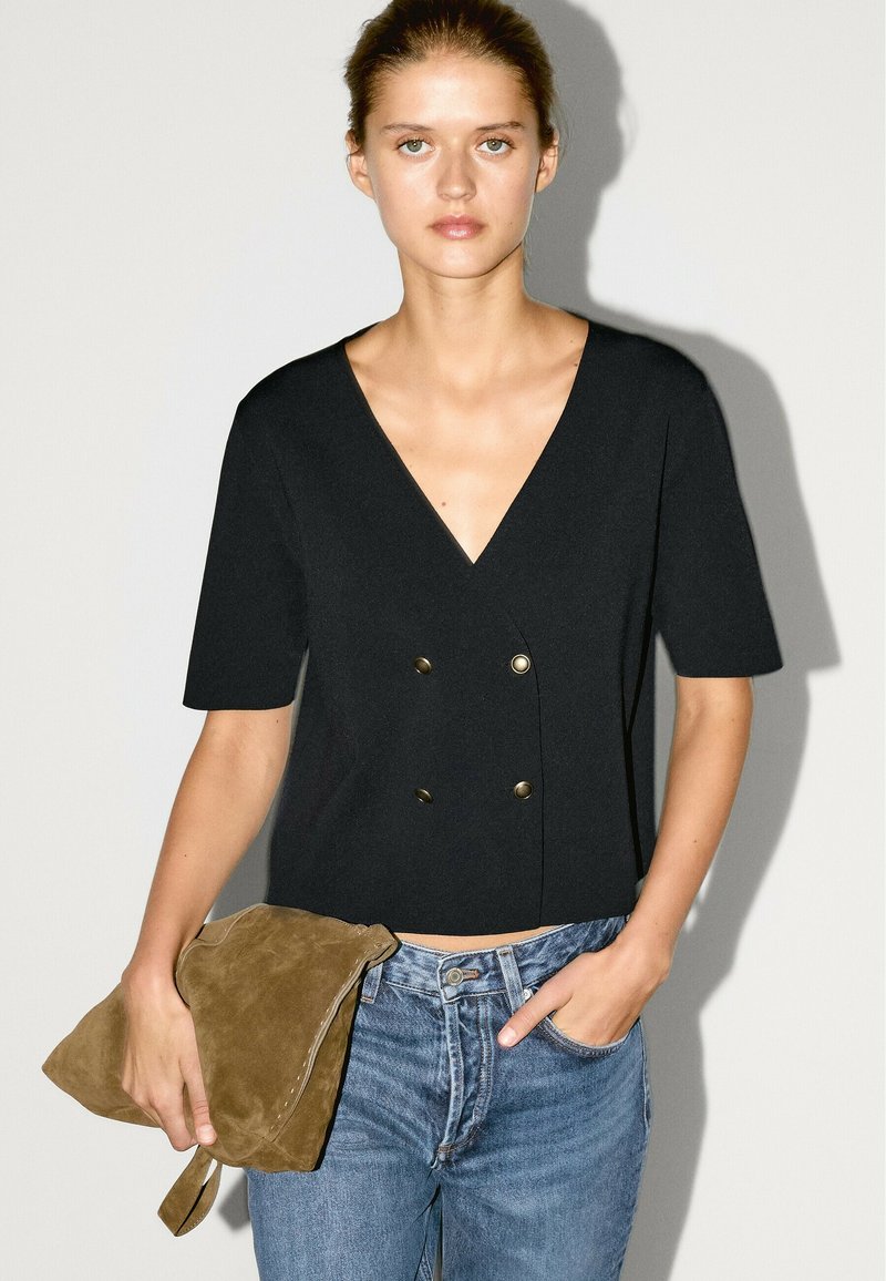 Schwarzes V-Ausschnitt-Oberteil mit kurzen Ärmeln und doppelten goldenen Knöpfen, kombiniert mit hellen Jeans. Das Model hält eine beige Wildleder-Clutch.