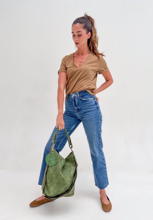 Frau in beigem Shirt und blauen Jeans, die eine große grüne Wildledertasche hält und vor einem schlichten weißen Hintergrund steht.