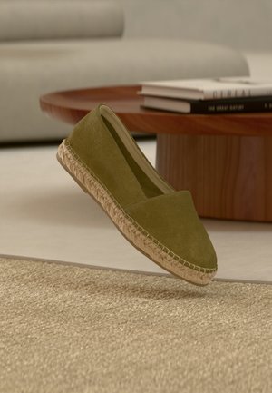 Espadrille en daim vert olive avec semelle en jute tressé flottant au-dessus d'un tapis beige tissé, d'une table basse en bois et d'une pile de livres en arrière-plan.