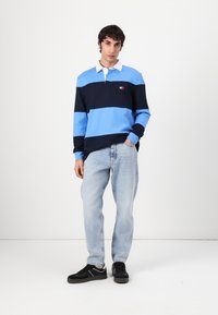 Blå og marineblå stripete langermet polo skjorte med hvit krage. Kombinert med lyseblå jeans og svarte joggesko. Enkel logo detalj på brystet.
