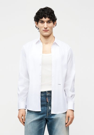 Jeune homme aux cheveux bouclés portant une chemise blanche ouverte sur un débardeur blanc et un jean bleu délavé, debout devant un fond uni.