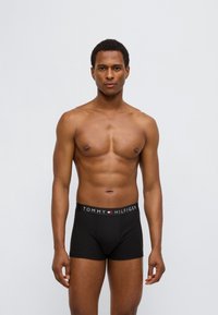 Μαύρα boxer briefs με λογότυπο στη μέση που αναγράφει "TOMMY HILFIGER" με λευκά γράμματα. Το μοντέλο είναι χωρίς μπλούζα, παρουσιάζοντας μυώδη σωματότυπο.