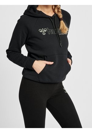 Kvinde iført sort hættetrøje og sorte leggings med logo, hænderne i forlommen, blondt hår i en sidefletning, grå baggrund.