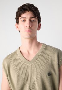 Colete de malha caqui com decote em V e mangas curtas, apresentando linhas verticais texturizadas e um pequeno logótipo no peito. Material suave.