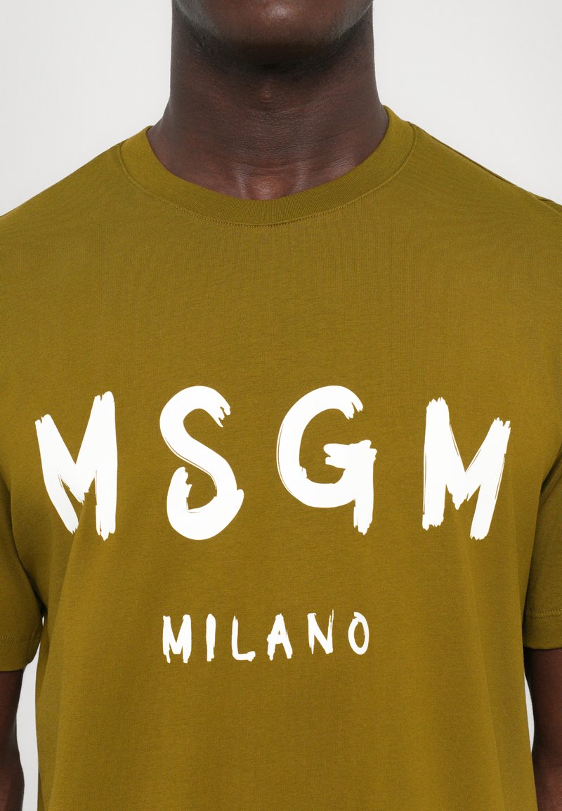 Olivgrön bomullst-shirt med fet vit text "MSGM" och "MILANO" i varierande storlekar. Rundad halsringning, korta ärmar. Mjuk textur.