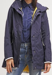Navy waterproof jacket with yellow interior, adjustable hood, and silver zippers. Features a relaxed fit and contrasting white cuffs.