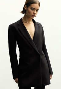 Schwarze Blazer mit tiefem V-Ausschnitt, strukturierten Schultern und einer einzelnen Knopfverschluss. Hergestellt aus glattem Stoff und ausgestattet mit zwei siden Taschen.