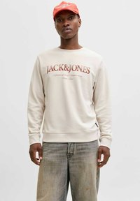 Sweat-shirt beige clair avec un logo rouge "JACK&JONES", col rond et poignets côtelés, associé à un jean gris délavé.