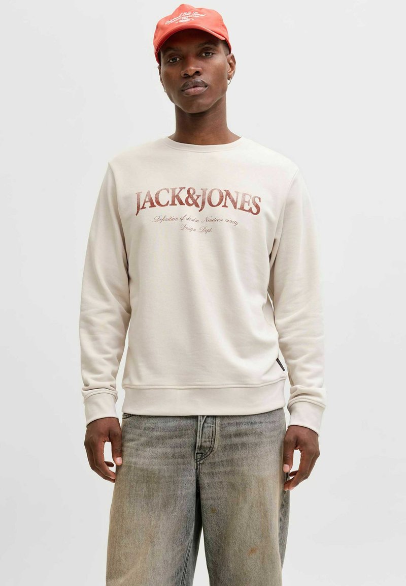 Sweat-shirt beige clair avec un logo rouge "JACK&JONES", col rond et poignets côtelés, associé à un jean gris délavé.