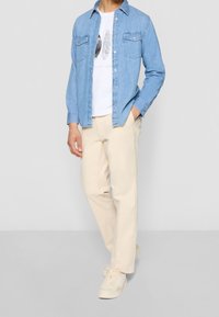 Lichtblauwe denim shirt over een witte graphic t-shirt, beige broek en witte sneakers. Het shirt heeft twee borstzakken en een knoopsluiting.