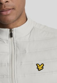 Veste blanche zippée avec une texture côtelée horizontale, arborant un petit logo d'aigle noir et jaune sur la poitrine. Tissu lisse et col montant.