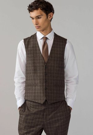 Gilet à carreaux marron en tissu texturé, avec un col à revers, des boutons sur le devant et des poches latérales, associé à une chemise blanche.