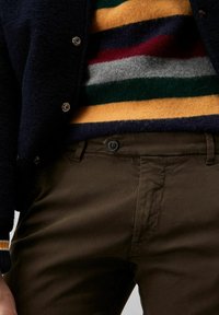 Pantaloni in cotone marrone con chiusura a bottone, abbinati a un maglione a strisce multicolori in tonalità di giallo, verde, rosso e grigio.
