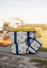 Trois sacs blancs et bleu marine à thème nautique avec des motifs marins et étiquetés "The Flat Lay Co. boating dept." placés à l'extérieur sur une rambarde en bois.