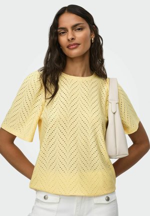 Kobieta z ciemnymi falującymi włosami, ubrana w żółtą, wzorzystą bluzkę z krótkim rękawem, białe spodnie, złote kolczyki-koła i nosząca białą torebkę na ramię.