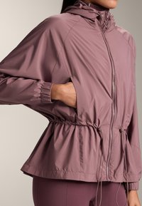 Lichte mauve windjack met een capuchon. Heeft elastische manchetten, een getailleerde taille en een rits aan de voorkant. Gladde textuur en losse pasvorm.