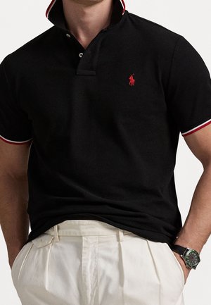 Man draagt een zwart poloshirt met rood logo en witte bies, ingestopt in een witte broek, handen in de zakken, met een horloge om de linkerpols.