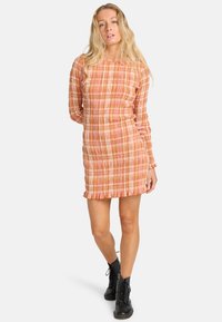 Robe à manches longues avec un design texturé et plissé dans des tons orange et beige, présentant une silhouette droite et un ourlet à volants. Associée à des bottes noires.