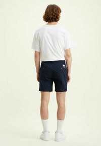 Laivastonsiniset ruudulliset shortsit, joissa on tasainen etuosa, sivutaskut ja takana brändilogo, yhdistettyinä yksiväriseen valkoiseen t-paitaan ja valkoisiin tenniskenkiin.