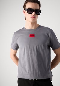 Grå bomulds t-shirt med en rød rektangulær lap, hvor der står "HUGO." Korte ærmer og en afslappet pasform, med en glat tekstur.
