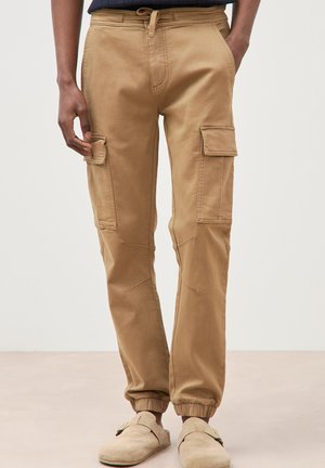 Man draagt beige cargobroek met elastische boorden en beige instapschoenen, staat met handen in de zakken op een neutrale achtergrond.