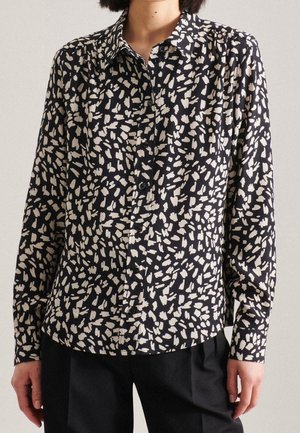 Personne portant une chemise noire à boutons avec un motif abstrait de coups de pinceau blancs et un pantalon noir, debout devant un fond uni.