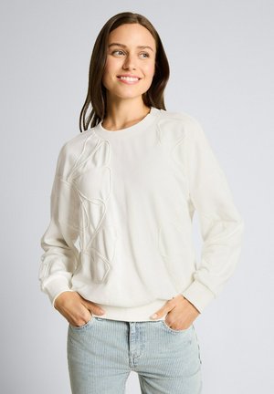 Młoda kobieta uśmiechająca się, ubrana w białą bluzę z wyszywanymi kwiatowymi wzorami oraz jasnoniebieskie jeansy w paski, ręce w kieszeniach, szare tło.