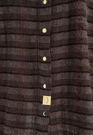 Brązowo-czarna kratka na tkaninie z rzędem okrągłych złotych guzików i małą beżową metką przedstawiającą zamknięte oko.