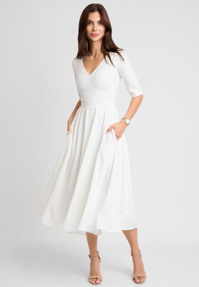 Biała sukienka do kolan z dopasowanym gorsem, dekoltem w kształcie litery V i krótkimi rękawami; ma rozkloszowany dół ze kieszeniami i gładką teksturą.