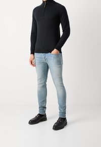 Schwarzer Zip-Kragen-Pullover, Slim-Fit hellblaue Jeans und schwarze Slipper mit goldenen Akzenten. Glatte Textur und taillierter Schnitt.