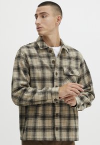 Chemise en flanelle beige avec un motif à carreaux noir et marron, dotée d'un boutonnage sur le devant, d'un col et d'une poche poitrine. Texture douce, design décontracté.