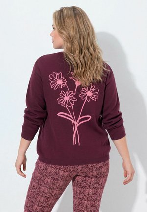 Vrouw met golvend blond haar die een donkerpaarse trui draagt met roze bloemdessin op de rug en roze broek met patroon.