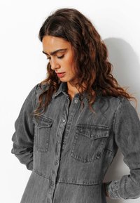Chemise en denim gris avec des manches longues, une fermeture à boutons sur le devant, deux poches poitrine, et une coupe légèrement décontractée avec une texture douce.