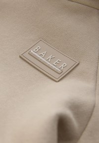 Gros plan sur un tissu beige avec un patch rectangulaire cousu indiquant "BAKER by Ted Baker" en lettres en relief.