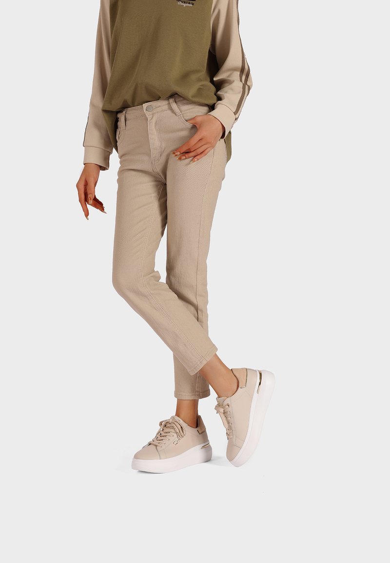 Pantaloni di cotone beige con superficie texturizzata, abbinati a sneaker beige con suola bianca e dettagli metallici. Maglia verde chiaro.