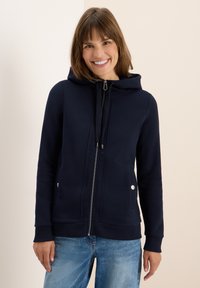 Haut à capuche zippé de couleur navy avec un cordon de serrage, des poches avant et des accents argentés. Fabriqué en tissu doux avec des poignets et un ourlet côtelés.