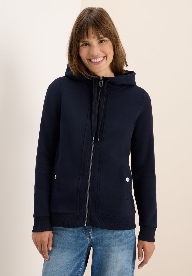 Haut à capuche zippé de couleur navy avec un cordon de serrage, des poches avant et des accents argentés. Fabriqué en tissu doux avec des poignets et un ourlet côtelés.