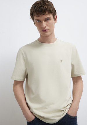 Junger Mann mit kurzen lockigen Haaren, der ein schlichtes hellbeiges Kurzarm-T-Shirt mit kleinem Besticktem Logo trägt, Hände in den Taschen.