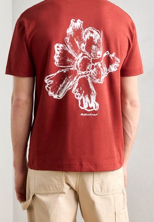 Mężczyzna w rdzawym t-shircie z dużym białym, abstrakcyjnym wzorem kwiatu i napisem „Marco Polo” na plecach, w połączeniu z beżowymi spodniami.