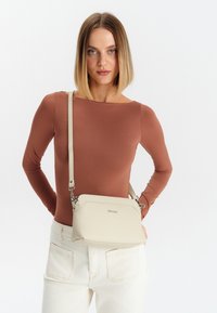 Borsa a tracolla in pelle beige testurizzata con design angolare, forma slim, tracolla regolabile e dettagli in metallo argentato.