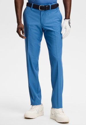 Man draagt blauwe op maat gemaakte broek, zwarte riem, witte golfhandschoen aan de rechterhand en witte sneakers, staand tegen een effen achtergrond.