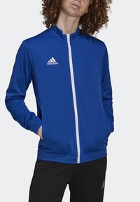 Blå sportjacka i slätt material, med hög krage, fullständig framdragkedja, vit Adidas-logga på bröstet och sidfickor.